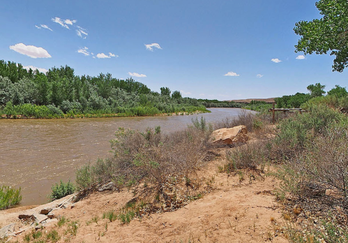 A calm river flows through a lush green landscape under a clear blue sky, with sandy banks and scattered rocks.